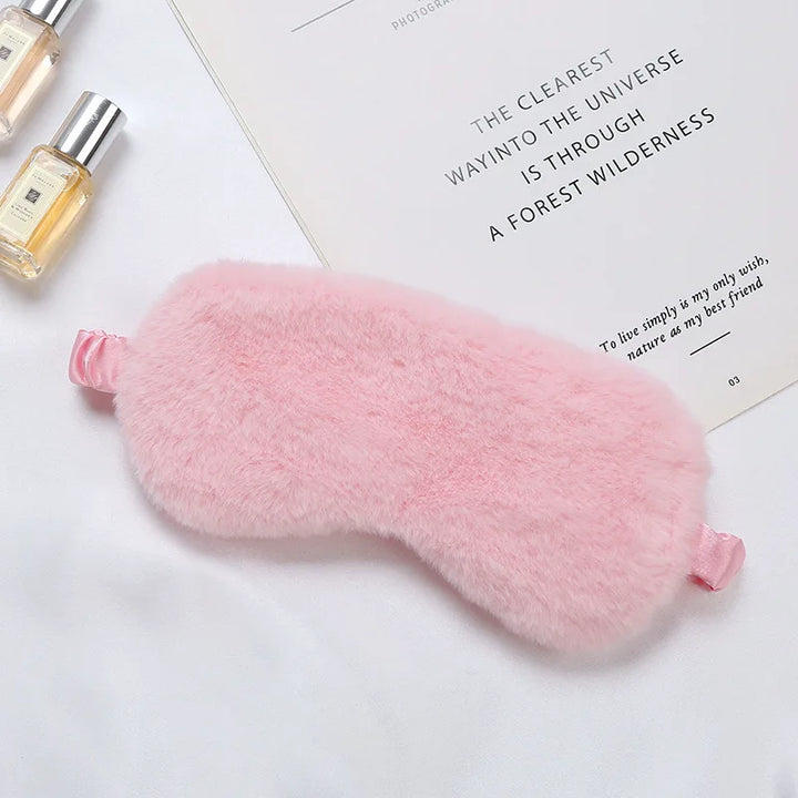 Mascherina per dormire in peluche rosa su sfondo bianco con flaconi di profumo e libro. Accessorio per il sonno, morbido e confortevole.