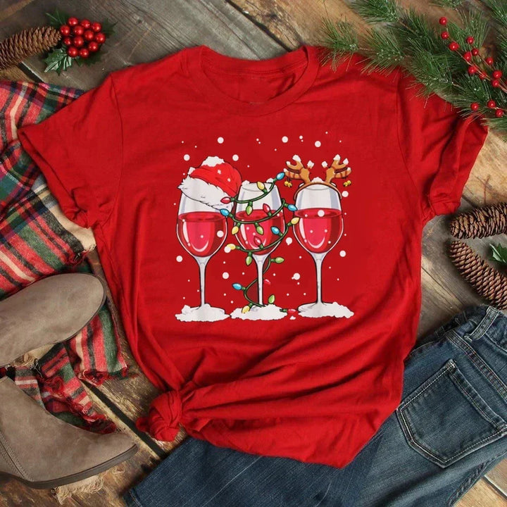 Maglietta rossa natalizia con tre bicchieri di vino decorati con cappello di Babbo Natale, luci e corna di renna, su sfondo di legno e pigne.
