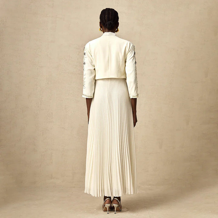 Donna di spalle con abito lungo plissettato bianco, giacca corta ricamata, orecchini dorati, su sfondo neutro. Moda elegante, abbigliamento femminile.