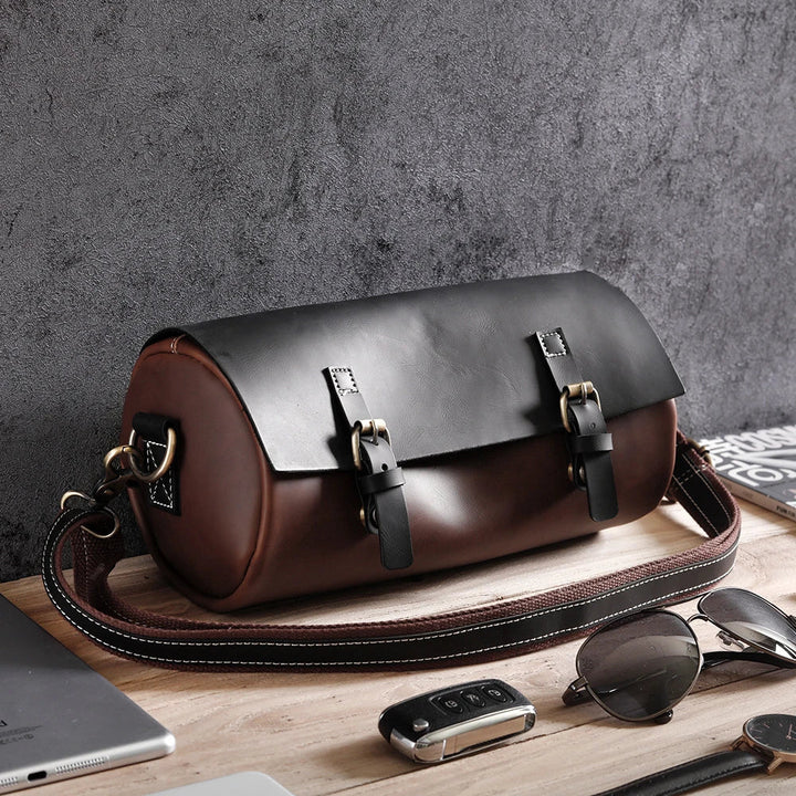 Borsa a tracolla in pelle marrone e nera su tavolo di legno, con fibbie metalliche. Accessorio moda elegante per uomo, ideale per viaggi e lavoro.