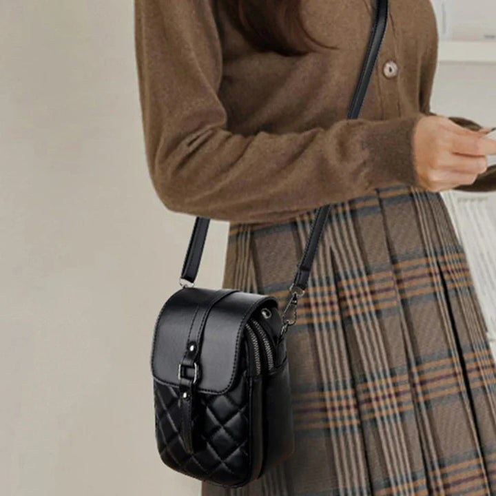 Borsa a tracolla nera trapuntata in pelle, indossata da donna con maglione marrone e gonna a quadri. Accessorio moda elegante e versatile.