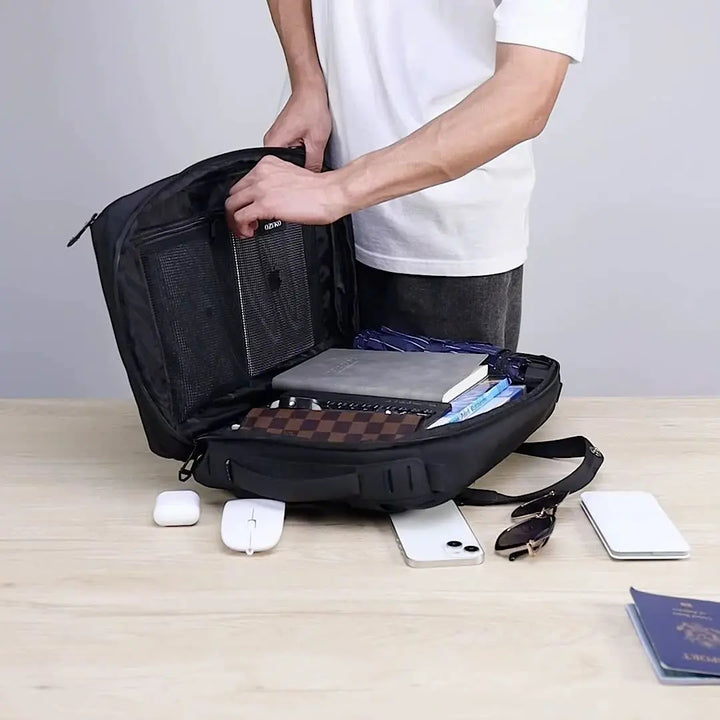Zaino nero aperto su tavolo, con libri, quaderni e accessori tecnologici. Uomo in maglietta bianca. Accessori: smartphone, mouse, auricolari.