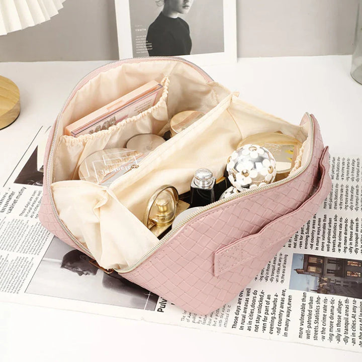 Astuccio da viaggio rosa in tessuto intrecciato, aperto, con scomparti organizzati contenenti cosmetici e accessori, su sfondo di giornale.