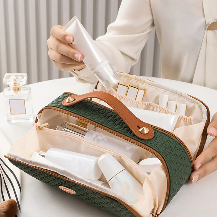 Borsa cosmetica verde con manico in pelle, contenente flaconi e tubetti di prodotti per la cura della pelle, su tavolo bianco accanto a profumo.