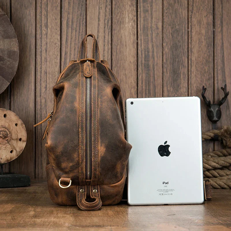 Zaino in pelle vintage marrone con zip verticale, accanto a un iPad, su sfondo in legno. Accessorio moda uomo, stile retrò, alta qualità.