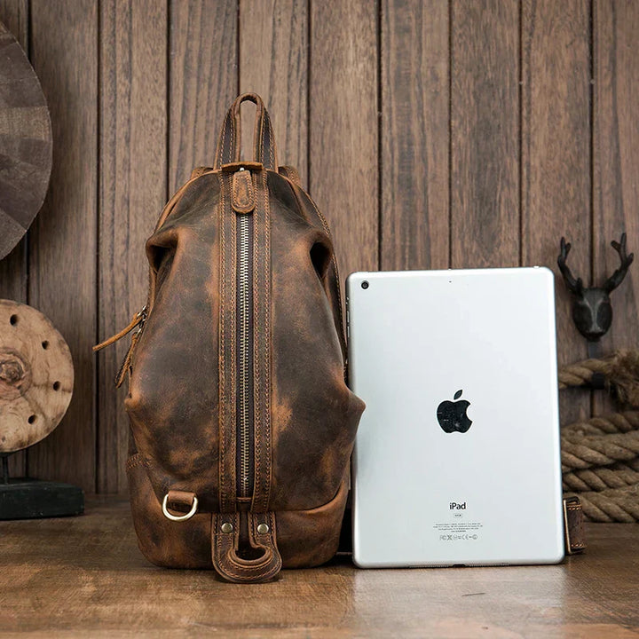 Zaino in pelle vintage marrone con zip verticale, accanto a un iPad, su sfondo in legno. Accessorio moda uomo, stile retrò, alta qualità.