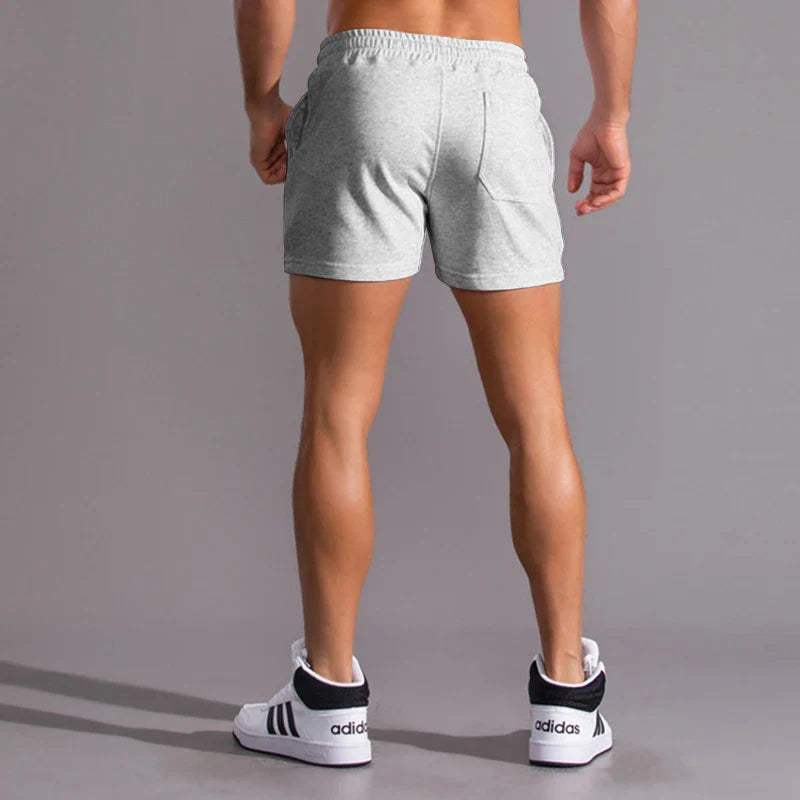 Shorts sportivi grigi da uomo, elasticizzati, con tasche laterali. Ideali per allenamento e fitness. Scarpe da ginnastica bianche con strisce nere.