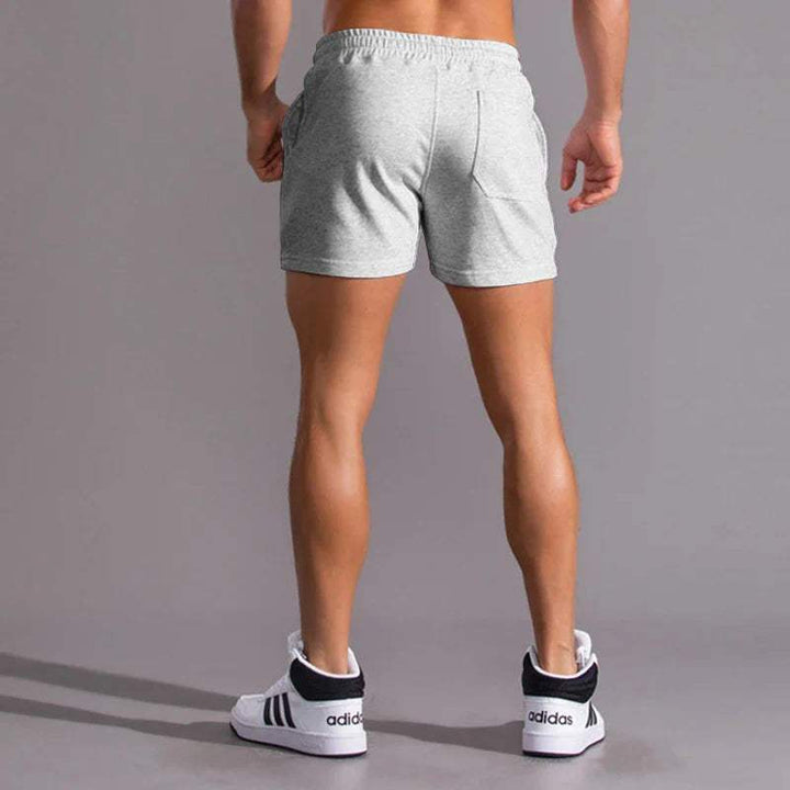 Shorts sportivi grigi da uomo, elasticizzati, con tasche laterali. Ideali per allenamento e fitness. Scarpe da ginnastica bianche con strisce nere.