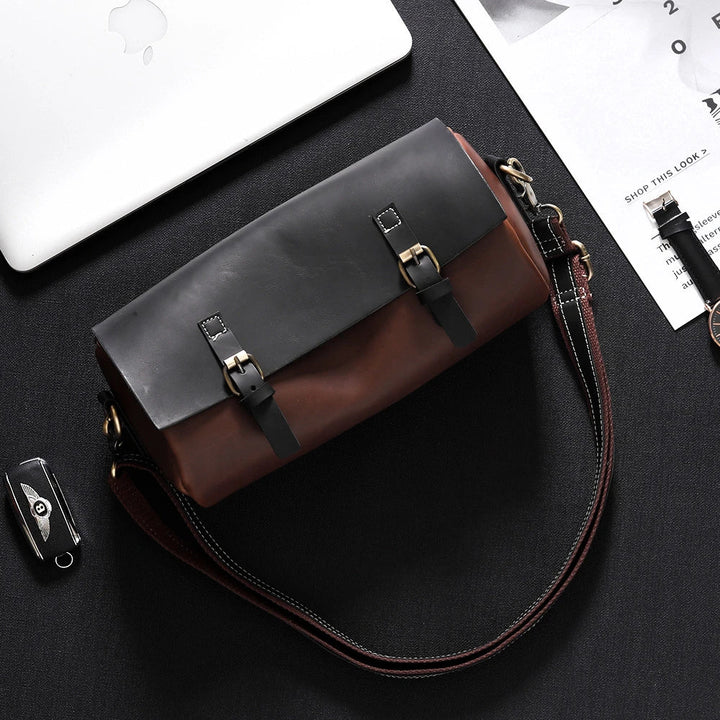Borsa a tracolla in pelle marrone e nera su sfondo nero, con fibbie metalliche. Accessorio moda elegante, ideale per uomo e donna.