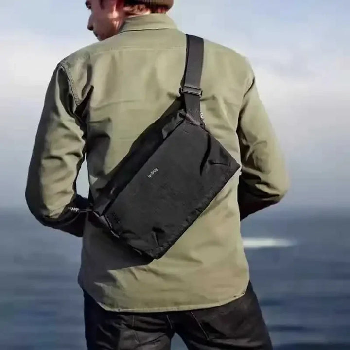 Uomo con borsa a tracolla nera all'aperto, giacca verde, vista mare. Accessorio moda maschile, borsa resistente, stile casual.