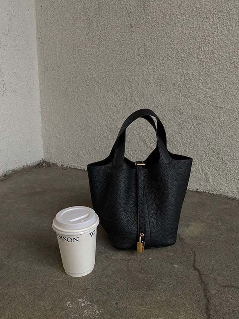 Borsa a secchiello nera in pelle con chiusura a zip, accanto a bicchiere di caffè da asporto su pavimento grigio. Accessorio moda elegante e moderno.