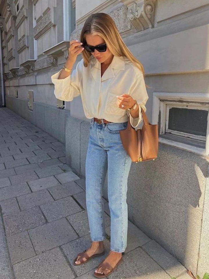 Donna con camicia bianca, jeans azzurri e sandali marroni, porta borsa in pelle marrone. Moda casual chic, street style estivo.