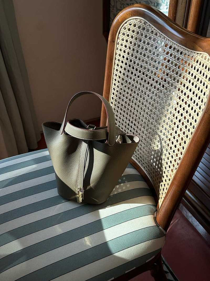 Borsa a secchiello in pelle verde oliva su sedia vintage con seduta a righe. Accessorio moda elegante, perfetto per look casual e chic.