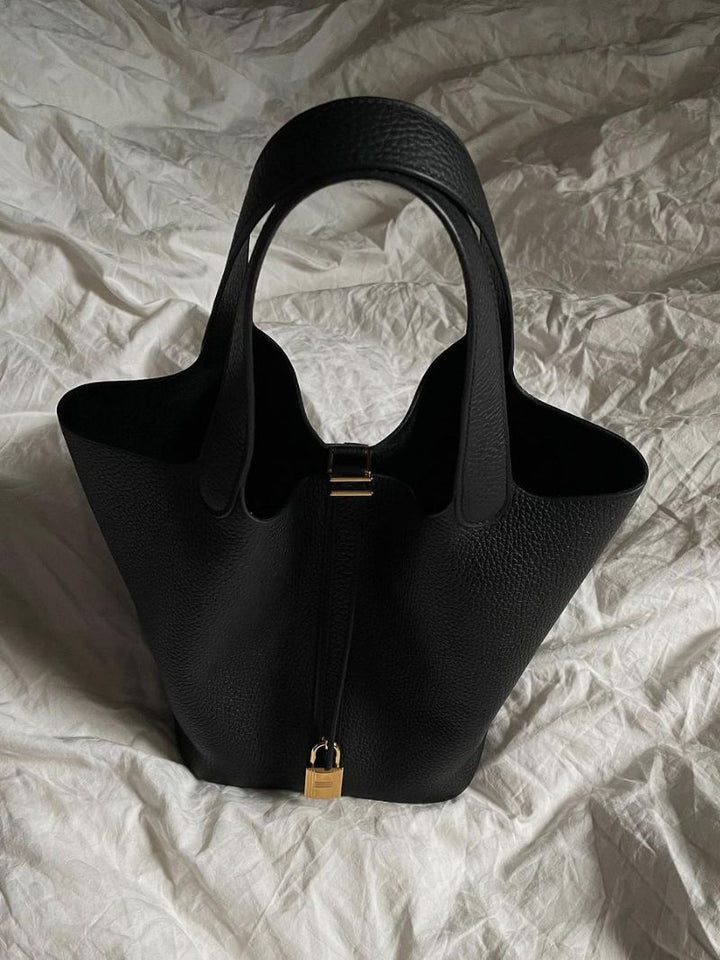 Borsa a mano nera in pelle su letto bianco, design elegante con chiusura dorata. Accessorio moda donna, stile sofisticato e moderno.