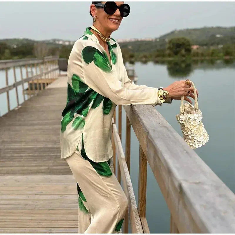 Donna elegante con occhiali da sole, indossa completo seta beige e verde, su passerella in legno vicino a lago, borsa a mano decorata.