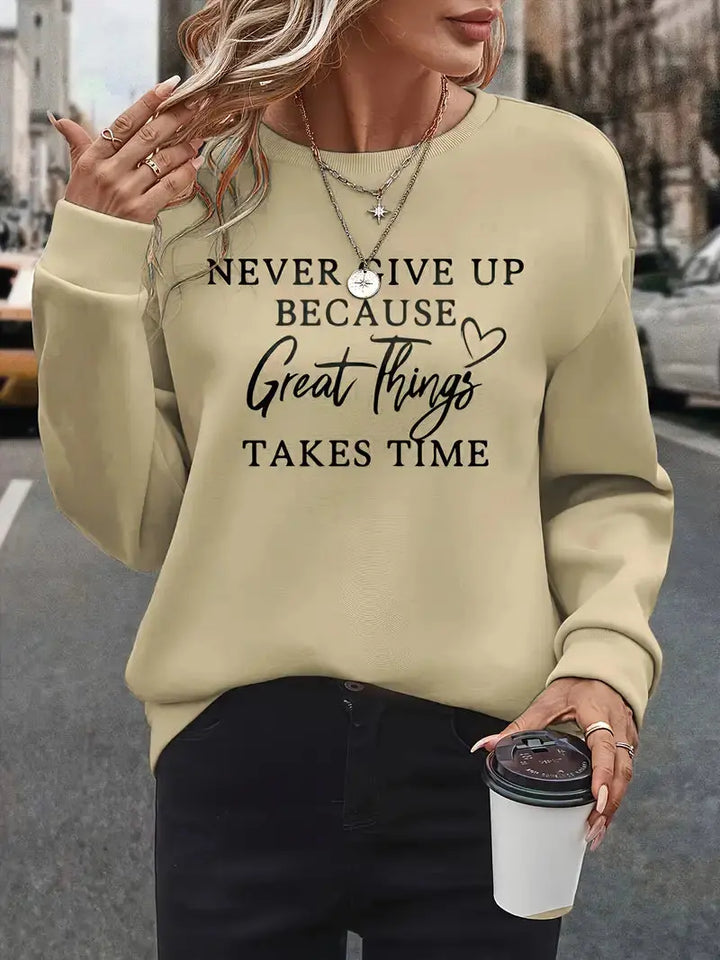 Donna con maglione beige con scritta motivazionale, collane a strati, pantaloni neri, tiene una tazza da caffè. Moda casual autunnale urbana.