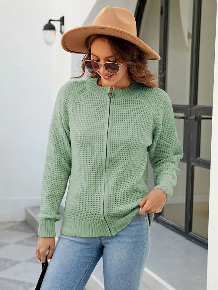 Donna con maglione verde a maglia, cappello beige e occhiali da sole, in piedi all'aperto. Moda autunnale, abbigliamento casual elegante.