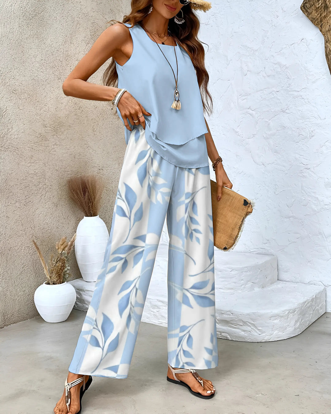 Donna con top azzurro e pantaloni a foglie bianche e blu, sandali neri, borsa di paglia, sfondo neutro. Moda estiva elegante e casual.