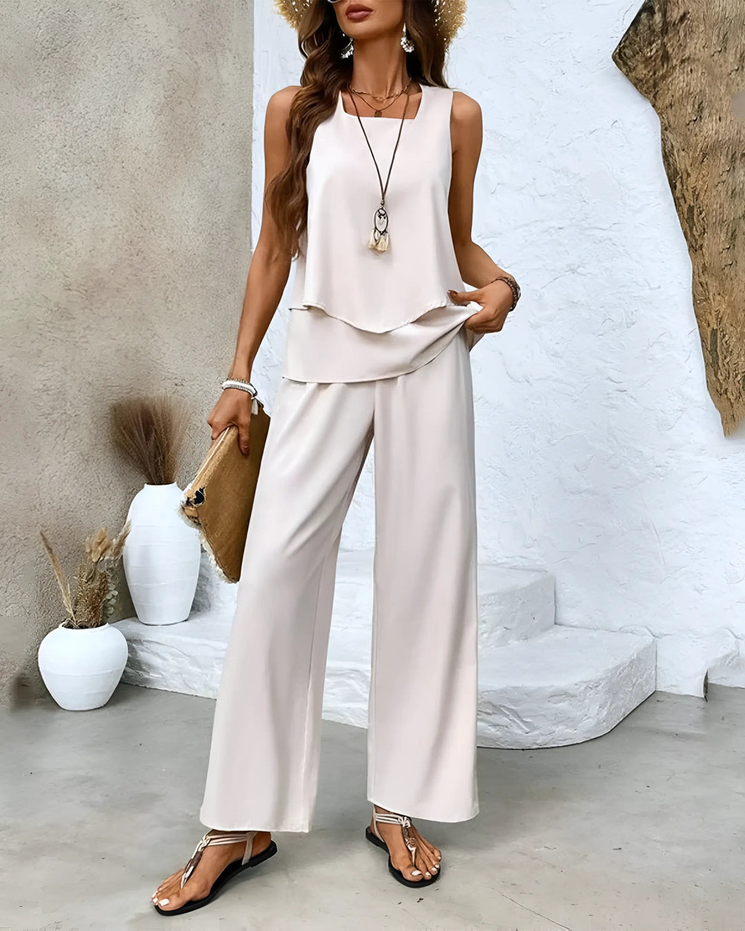Donna in elegante completo estivo bianco, top senza maniche e pantaloni larghi, accessoriata con cappello di paglia e borsa a mano, stile casual chic.