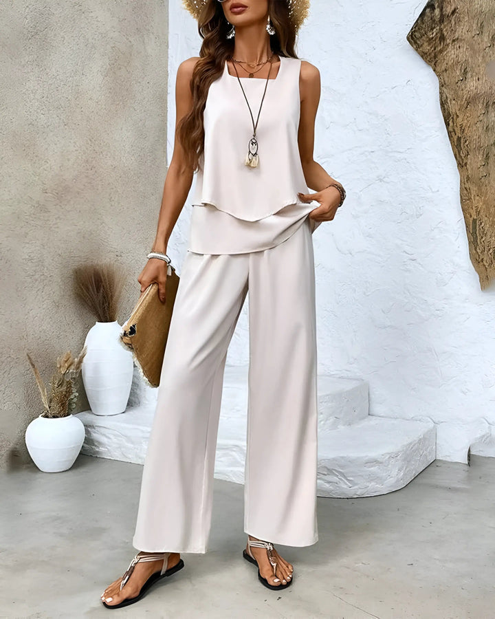 Donna in elegante completo estivo bianco, top senza maniche e pantaloni larghi, accessoriata con cappello di paglia e borsa a mano, stile casual chic.