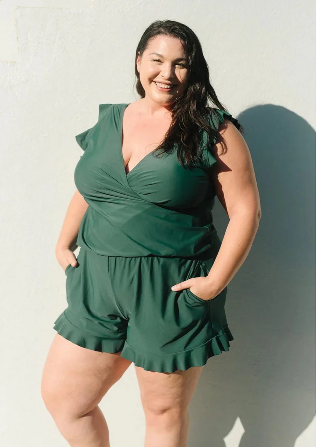 Donna sorridente in tuta verde, moda taglie forti, posa contro un muro bianco. Abbigliamento estivo elegante, stile casual chic.