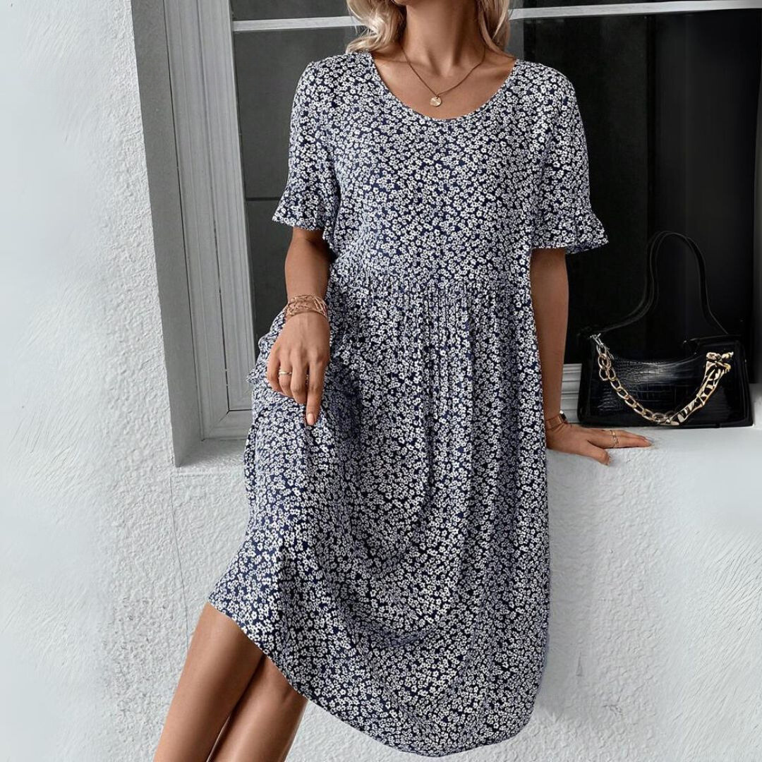 Donna con abito estivo floreale blu, maniche corte, accanto a borsa nera con catena dorata. Moda femminile casual elegante.