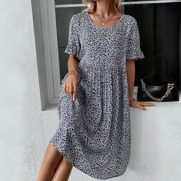 Donna con abito estivo floreale blu, maniche corte, accanto a borsa nera con catena dorata. Moda femminile casual elegante.