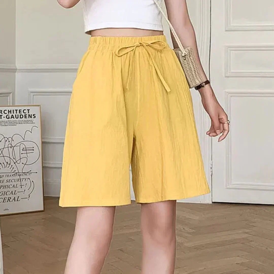 Donna con pantaloncini gialli a vita alta, top bianco corto e borsa a tracolla beige. Stile estivo casual, moda femminile trendy.