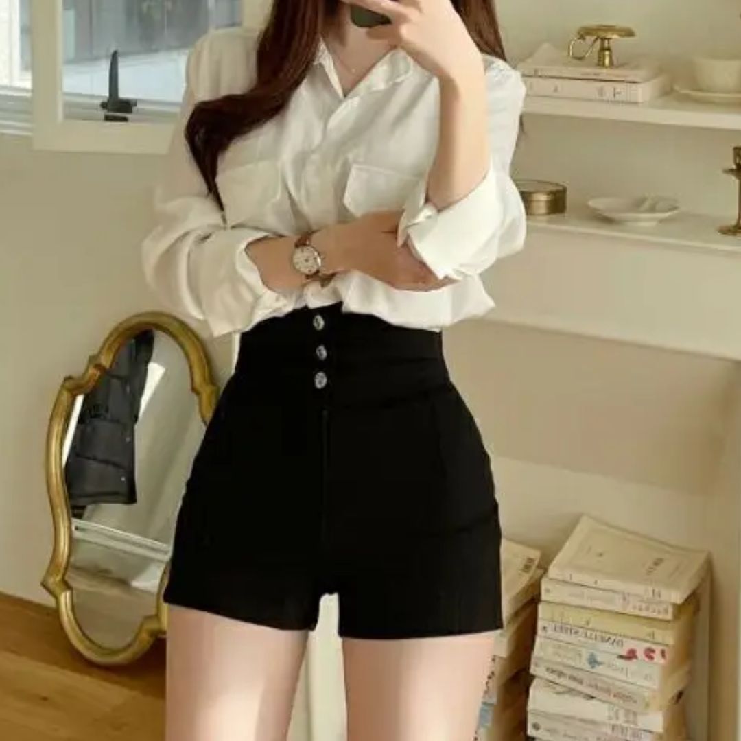 Donna in camicia bianca e pantaloncini neri a vita alta, in una stanza con specchio dorato e libri. Look elegante e moderno per moda casual.