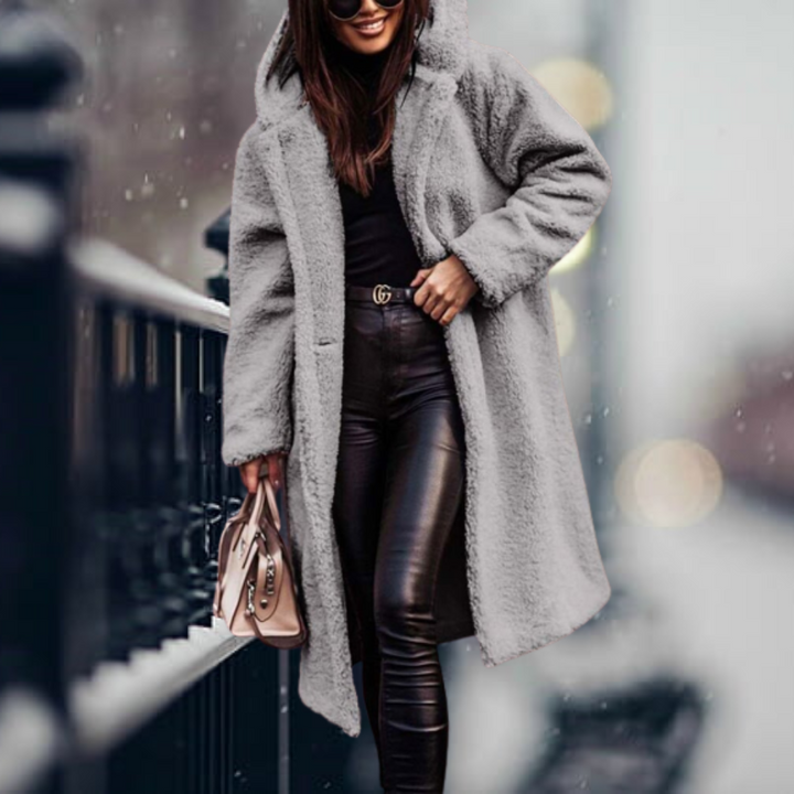 Donna con cappotto in pelliccia sintetica grigia, pantaloni in pelle nera, borsa beige, sorridente in strada innevata. Moda invernale elegante.