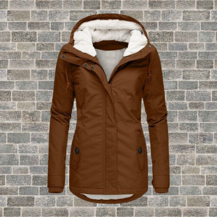 Giacca invernale da donna marrone con cappuccio foderato in pelliccia sintetica, chiusura a zip, tasche frontali, su sfondo di mattoni grigi.