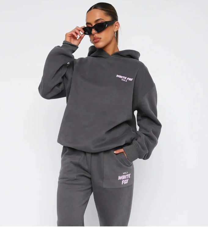 Donna con occhiali da sole indossa tuta sportiva grigia con cappuccio, marchio White Fox, moda casual, abbigliamento comodo, stile urbano.