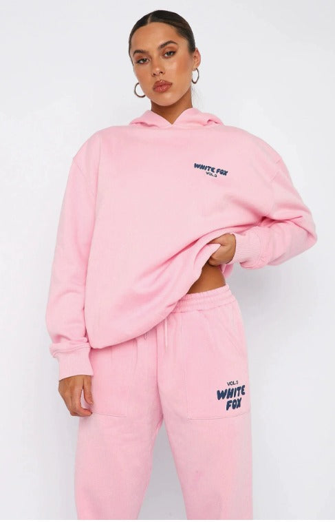 Donna con felpa e pantaloni rosa White Fox, logo visibile, posa casual. Abbigliamento sportivo alla moda, stile streetwear femminile.