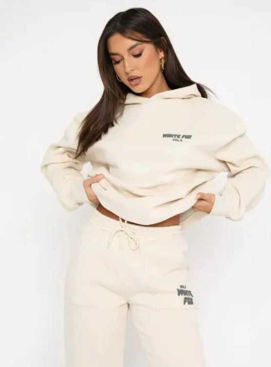 Modella indossa tuta sportiva beige con cappuccio, logo "White Fox" visibile. Abbigliamento casual donna, moda autunno, stile confortevole.