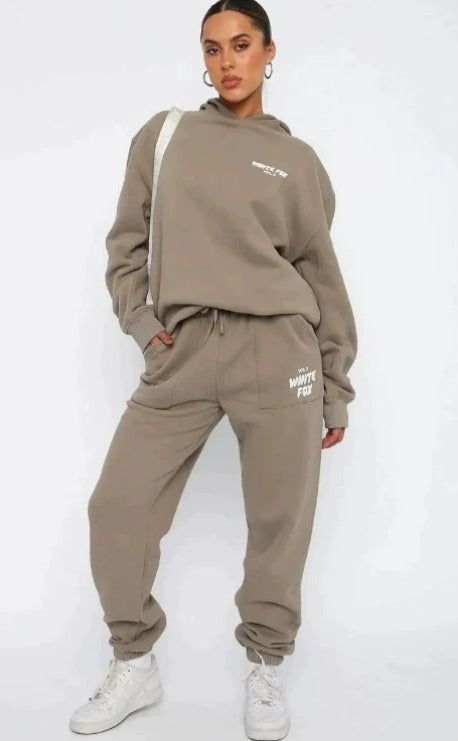 Donna in tuta sportiva beige, felpa con cappuccio e pantaloni abbinati, scarpe da ginnastica bianche. Moda casual, abbigliamento comodo.