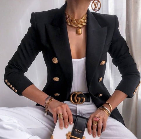 Donna elegante in blazer nero con bottoni dorati, top bianco, pantaloni bianchi. Accessori di lusso: collana, cintura, anelli, borsa Prada.