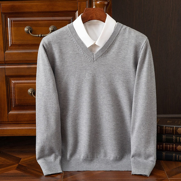 Maglione grigio in lana con scollo a V su camicia bianca, appeso su appendiabiti di legno, davanti a cassetti in legno scuro.