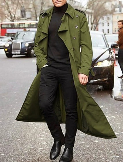 Uomo con trench verde oliva, maglione nero e pantaloni scuri, cammina in strada cittadina. Moda autunnale, stile urbano, cappotto elegante.