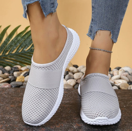 Scarpe slip-on grigie da donna, tessuto traspirante, suola bianca antiscivolo, indossate con jeans strappati, sfondo con pietre decorative.