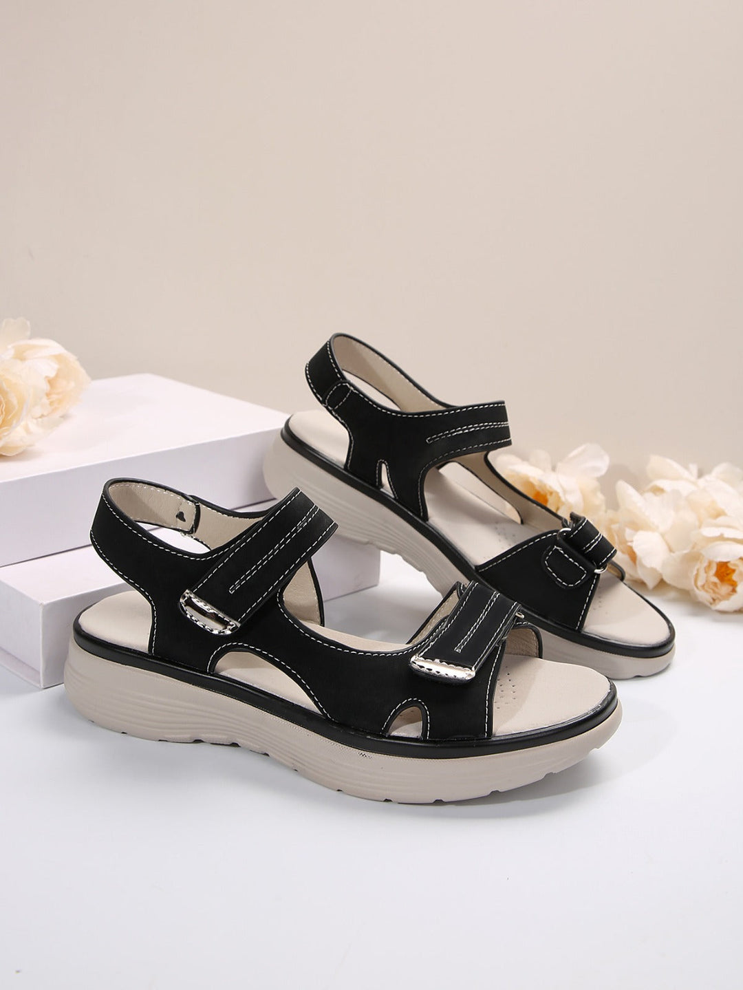 Sandali neri da donna con suola spessa beige, design sportivo, chiusura a strappo, su sfondo neutro con fiori bianchi.