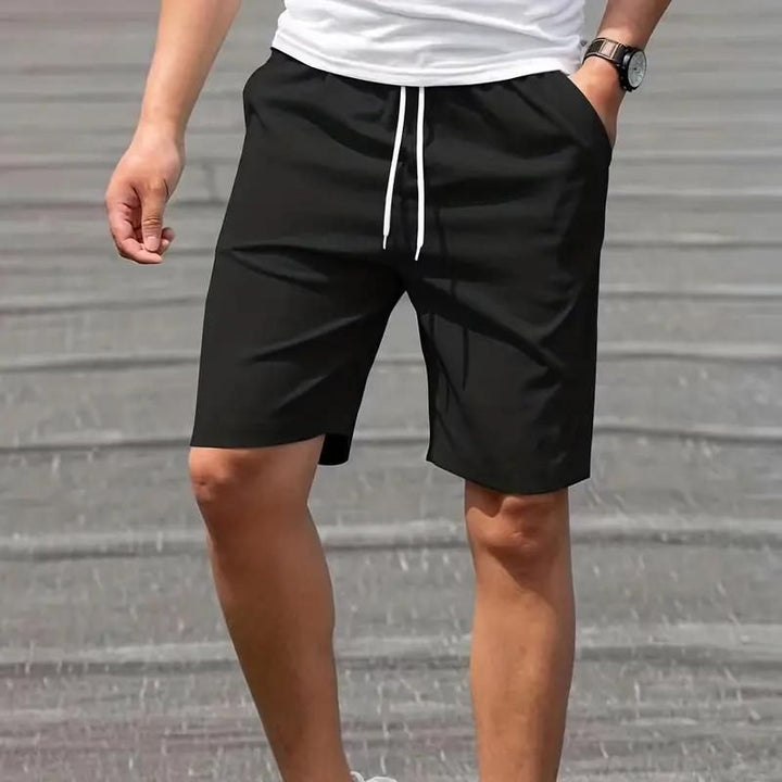 Uomo con pantaloncini neri casual, cordoncino bianco, maglietta bianca, orologio da polso, su sfondo grigio. Moda estiva maschile.