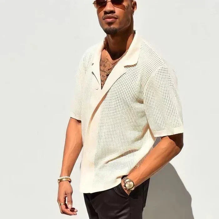 Uomo con camicia a maniche corte traforata bianca, occhiali da sole, orologio dorato, posa contro un muro bianco. Moda estiva casual elegante.