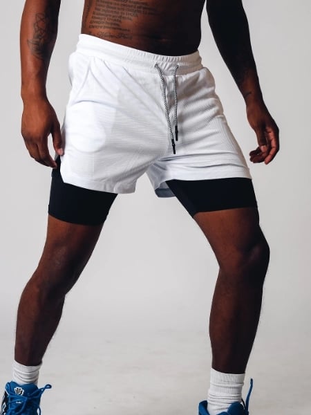 Uomo in pantaloncini sportivi bianchi con leggings neri, scarpe da ginnastica blu. Abbigliamento fitness maschile, moda sportiva, allenamento.
