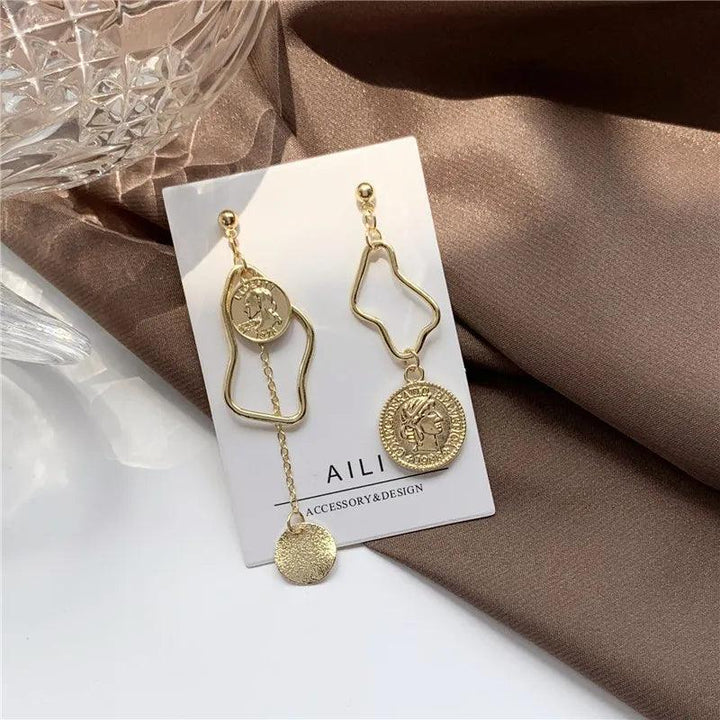 Orecchini pendenti asimmetrici in oro con design geometrico e monete, su sfondo marrone. Accessori moda eleganti per donna.