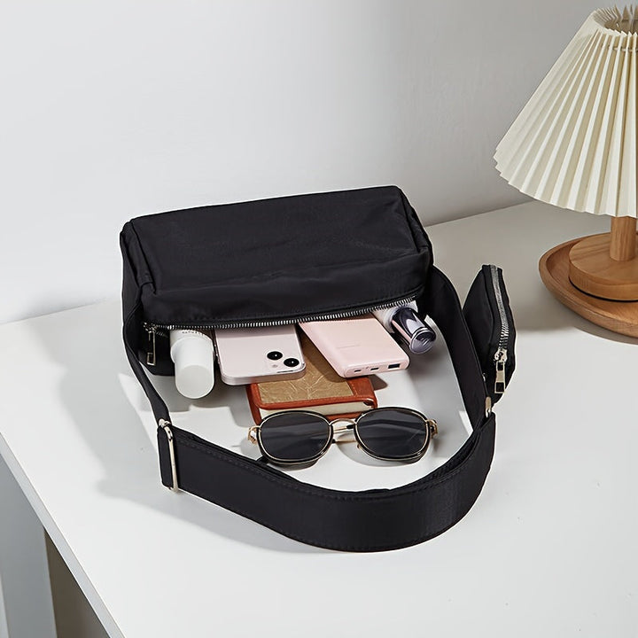 Borsa a tracolla nera su tavolo bianco con occhiali da sole, smartphone, rossetto e lampada. Accessori moda, eleganza, praticità quotidiana.