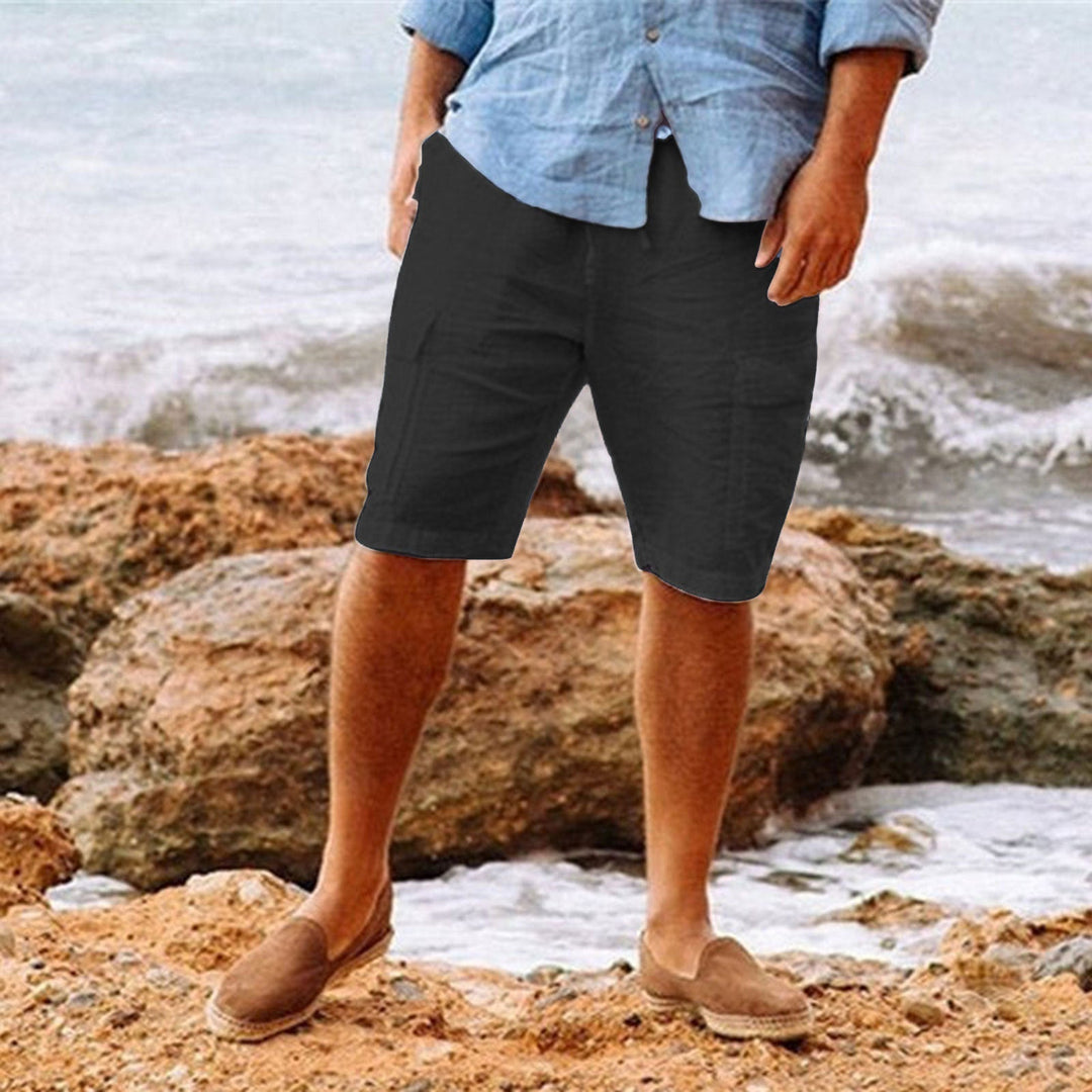 Uomo in pantaloncini neri e camicia azzurra su spiaggia rocciosa, indossa mocassini marroni. Moda estiva casual, abbigliamento maschile elegante.
