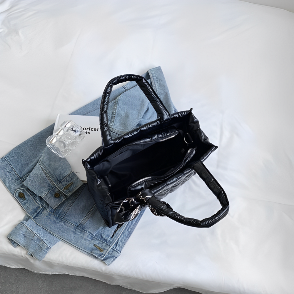 Borsa nera in pelle su letto bianco con giacca di jeans e libro. Accessori moda donna, stile casual chic.