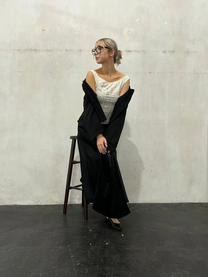 Donna elegante con occhiali, abito bianco e nero, appoggiata a uno sgabello su sfondo neutro. Moda contemporanea, stile minimalista.