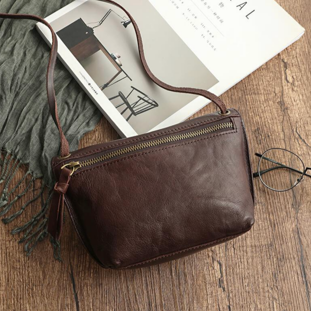 Borsa a tracolla in pelle marrone su tavolo di legno, accanto a occhiali e rivista. Design elegante, chiusura con zip, accessorio moda donna.