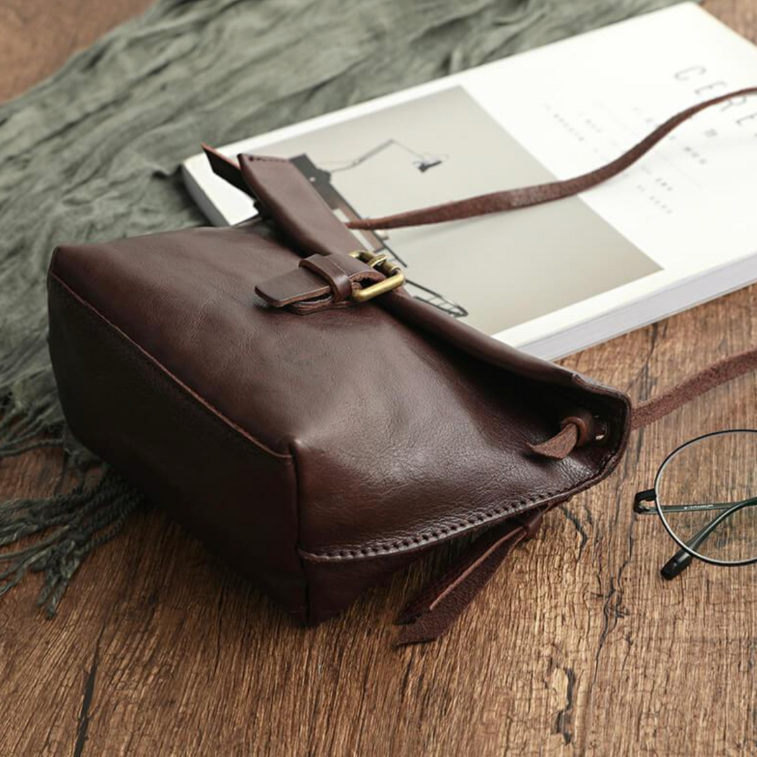 Borsa a tracolla in pelle marrone su tavolo di legno, accanto a libro e occhiali. Design elegante, ideale per moda e accessori di lusso.